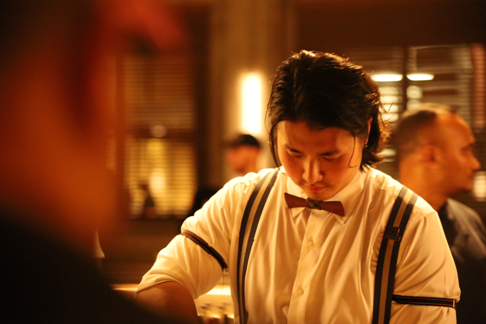Bartender in a white shirt, bow tie and suspenders working behind a warmly lit bar