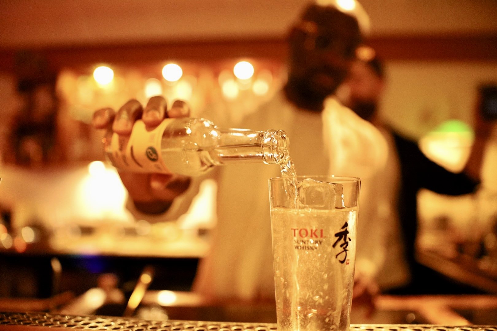 Bartender pouring soda into a highball glass over ice in warm candlelight